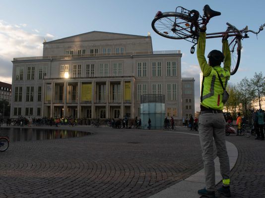 Mensch stemmt Fahrrad über den Kopf, im Hintergrund das Leipziger Opernhaus