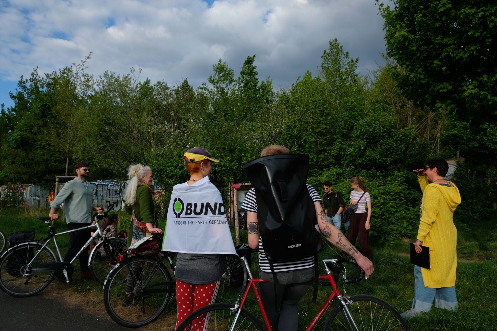 Es sind mehrere Menschen von hinten zu sehen. Eine Person trägt ein Banner mit der Auffschrift "BUND" auf dem Rücken. Die Menschen stehen von Bäumen und haben Fahrräder dabei.