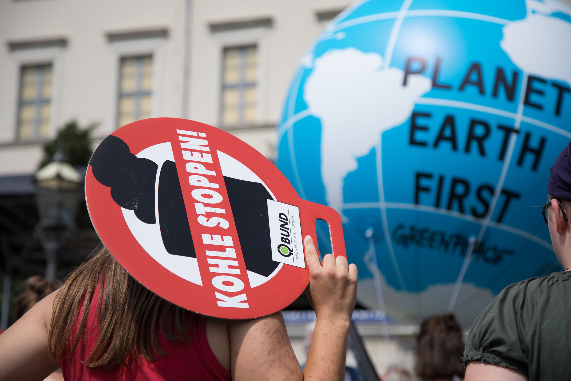 Frau von hinten, hält Schild auf dem steht: "Kohle stoppen"; im Hintergrund aufgeblasener Erdball auf dem steht: "Planet Earth First, Greenpeace"