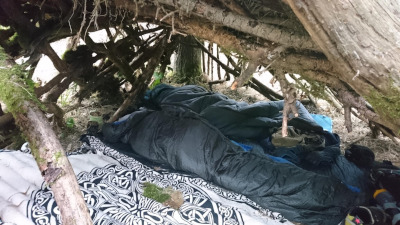 in einer selbstgebauten Hütte aus Ästen und Moos liegen zwei Schlafsäcke, in denen Menschen schlafen. 