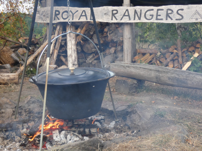 Dreibein Kocher steht über dem Feuer. Dahinter ist "Royal Rangers" auf einer Holzleiste zu lesen.