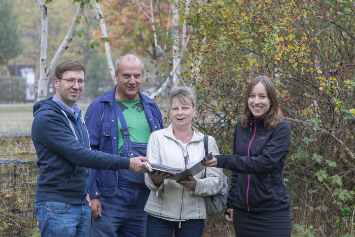 Vier Menschen halten ein Dokument in der Hand, im Hintergrund Bäume, Büsche, eine Mauer und ein Park