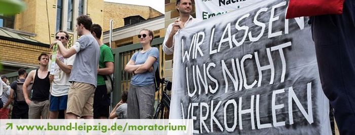 Menschen demonstrieren vor Stadtwerken, zwei halten ein Banner auf dem steht: "Wir lassen uns nicht verkohlen"; unten links steht Link: www.bund-leipzig.de/moratorium