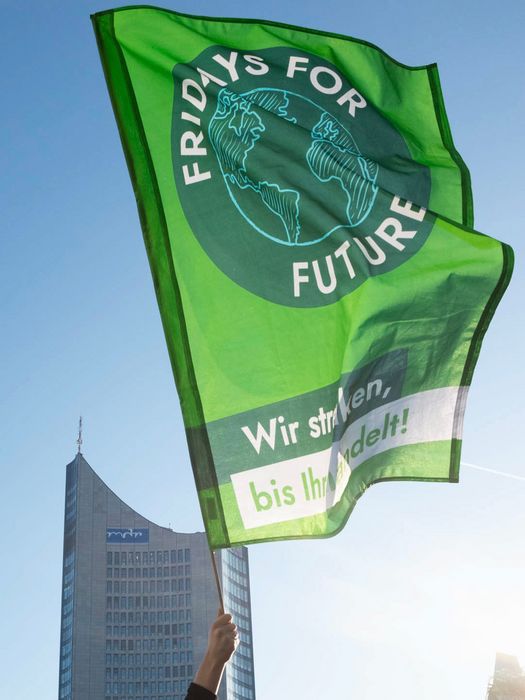 Eine Hand hält eine Fridays For Future Fahne hoch. Im Hintergrund befindet sich die Spitze des MDR Turms. Der Himmel ist blau und wolkenlos.