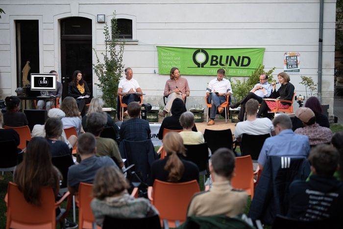 Menschen sitzen draußen vor einem Gebäude auf einem Podium und diskutieren. Vor Ihnen sitzen Menschen und schauen zu. 