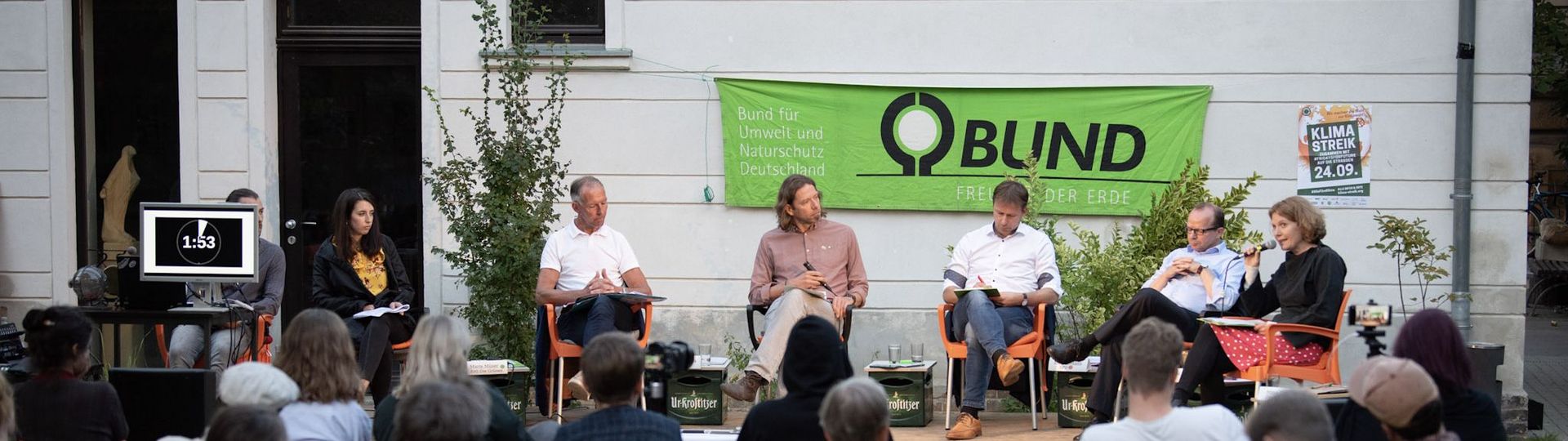 Menschen sitzen draußen vor einem Gebäude auf einem Podium und diskutieren. Vor Ihnen sitzen Menschen und schauen zu. 
