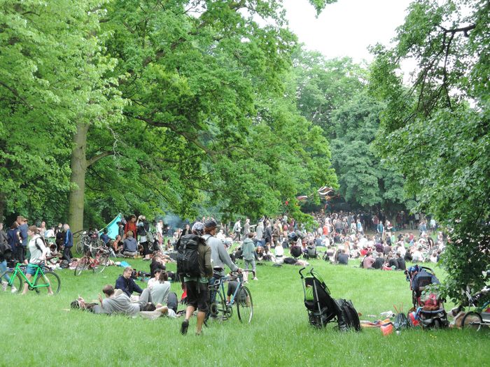 viele Menschen im Park, einige sitzen auf Decken, im Vordergrund schiebt jemand ein Fahrrad