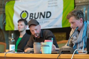Drei der neuen Mehrwegbecher im Vordergrund auf dem Podium, dahinter die Projektpartner