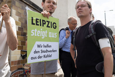 zwei Männer im Vordergrund; einer hält ein Schild auf dem steht: "Leipzig steigt aus. Wir helfen den Stadtwerken aus der Kohle!"