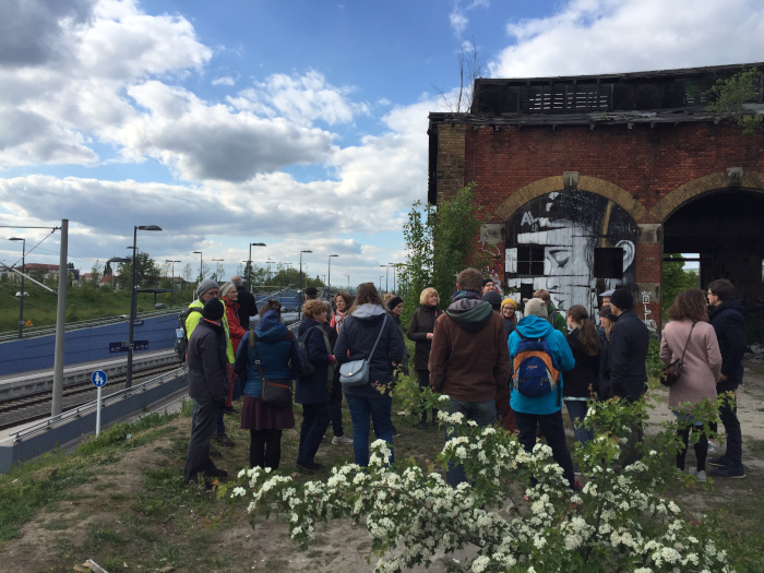 eine Gruppe von Menschen steht inmitten von Bahngleisen, Brachgelände und einem alten Ziegelsteingebäude, eine Person hält einen Vortrag zu den Örtlichkeiten