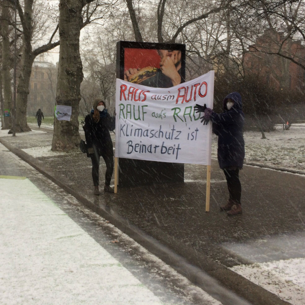 zwei Personen stehen am Rand einer Pop-up Bikelane und halten ein Banner hoch mit dem Text: "Raus aus dem Auto, rauf aufs Rad, Klimaschutz ist Beinarbeit"