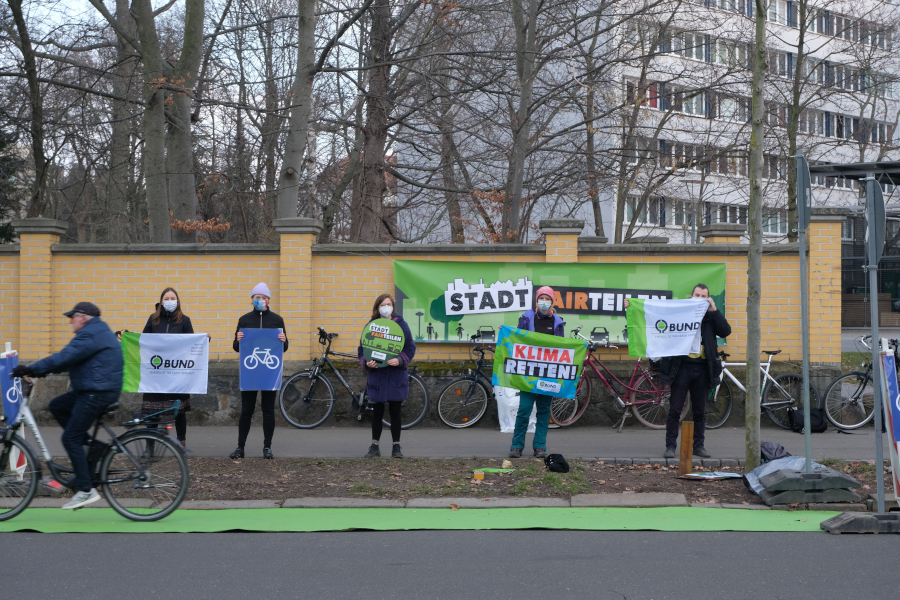 5 Personen stehen am Rand einer Pop-up Bikelane und halten Schilder und Transparente hoch zum Thema "Stadt Fairteilen" und "Klima retten".